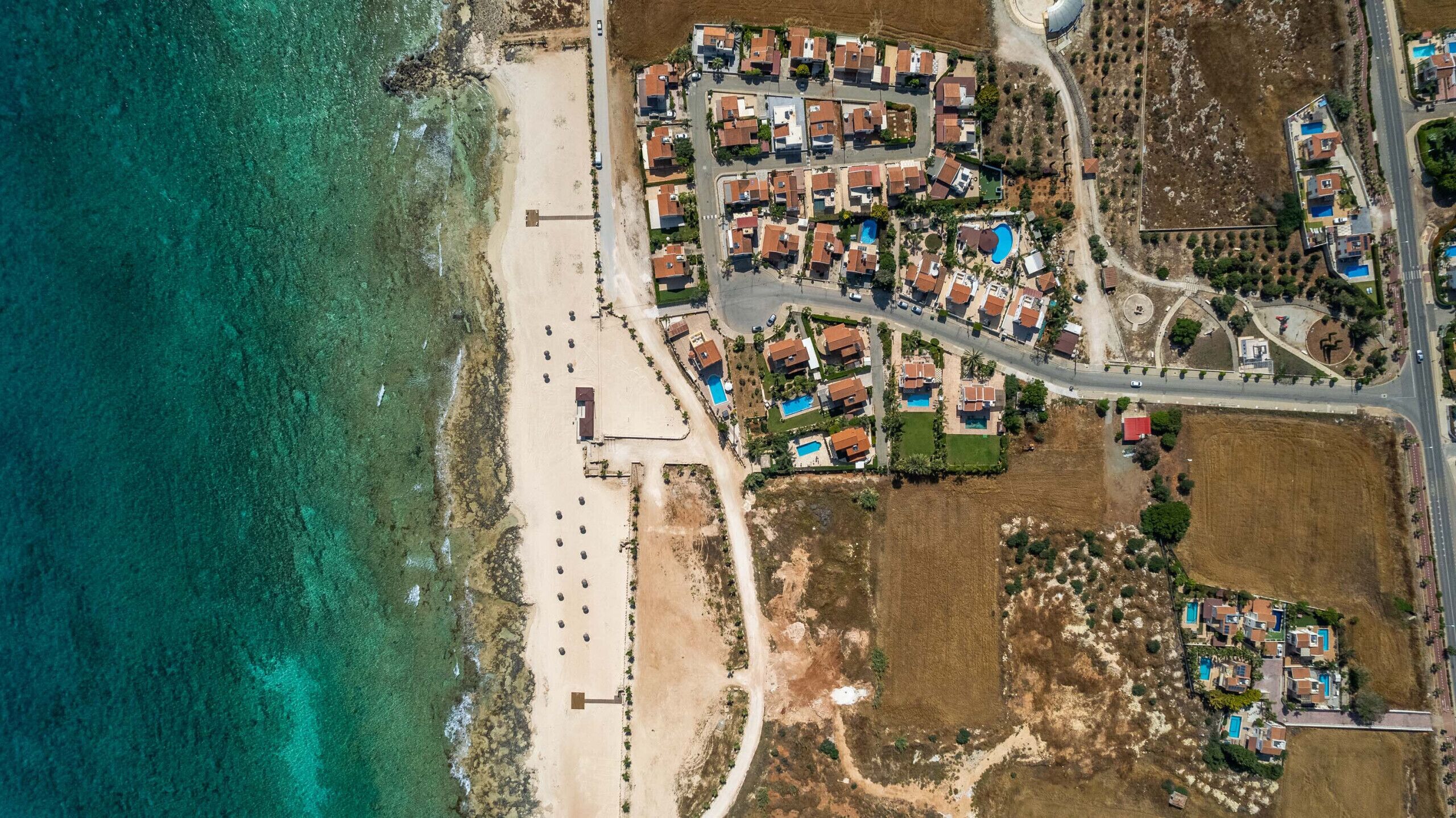 An aerial view of a coastal residential area with houses, a beach, and a road.