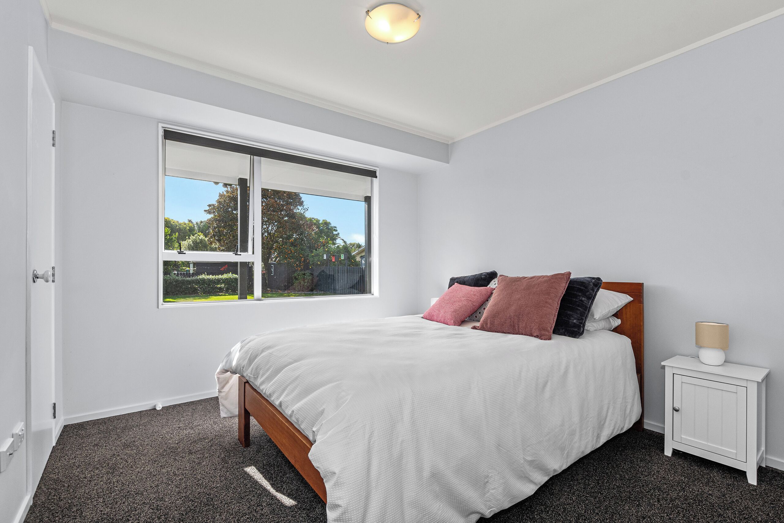 A bedroom with a large window, a bed with white bedding, and a nightstand with a lamp.