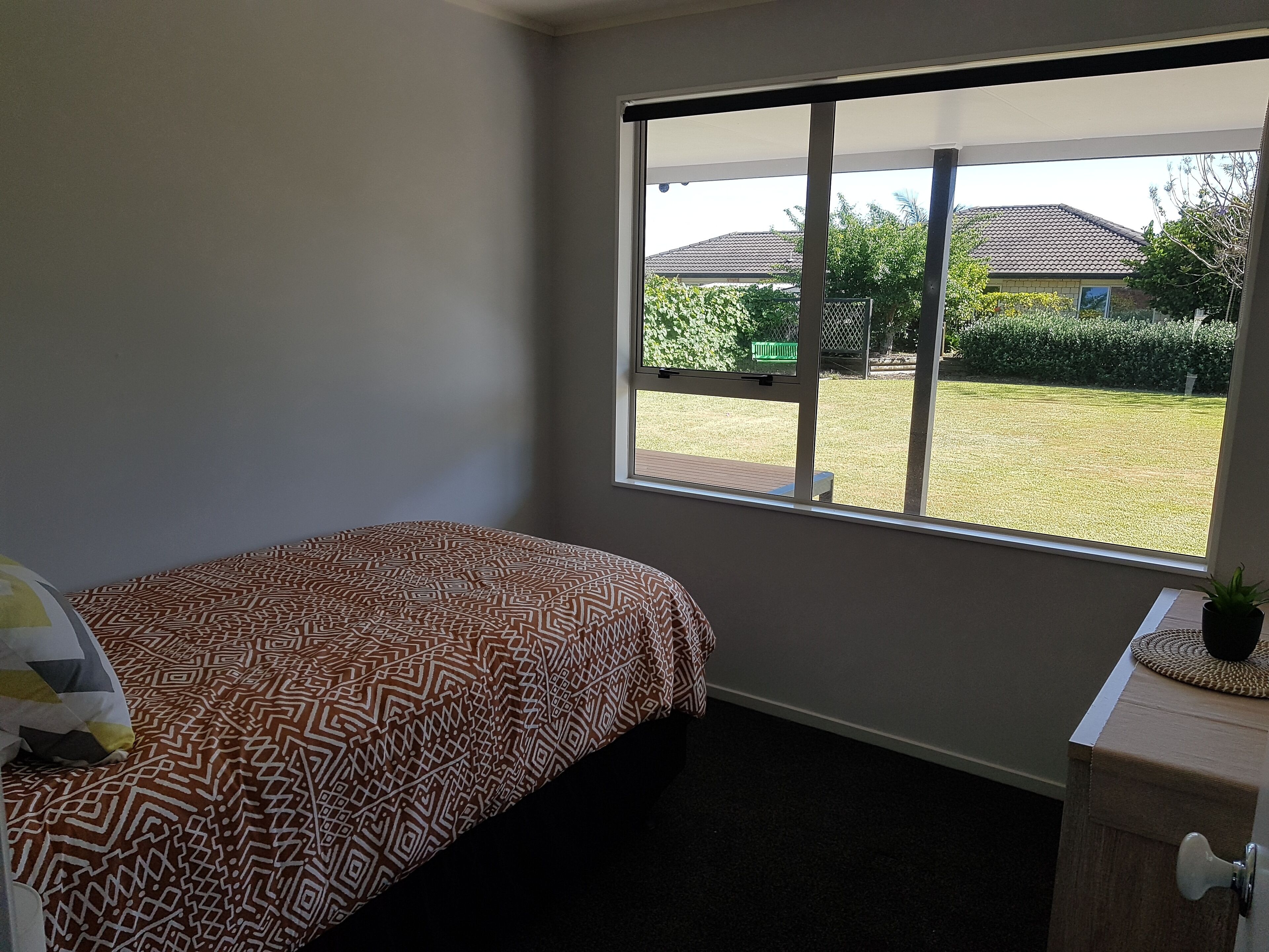A bedroom with a large window, a bed with patterned bedding, a bedside table with a plant, and a view of a lawn and houses outside.