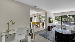 A modern living room with a glass desk, a white chair, a black patterned sofa, and a large window with a view of greenery.