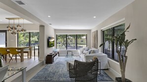 A modern living room with a sofa, dining table, and chairs, large windows, and a potted plant.