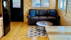 Ein gemütliches Blockhaus-Interieur mit Holzausstattung, einem schwarzen Kühlschrank, einem Ledersofa mit blauen Kissen, einem kleinen runden Tisch und einem Holzessttisch.
