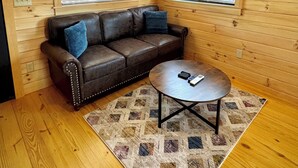 Ein gemütliches Zimmer mit Holzwänden, einem Ledersofa, einem runden Couchtisch und zwei Fenstern mit Jalousien.