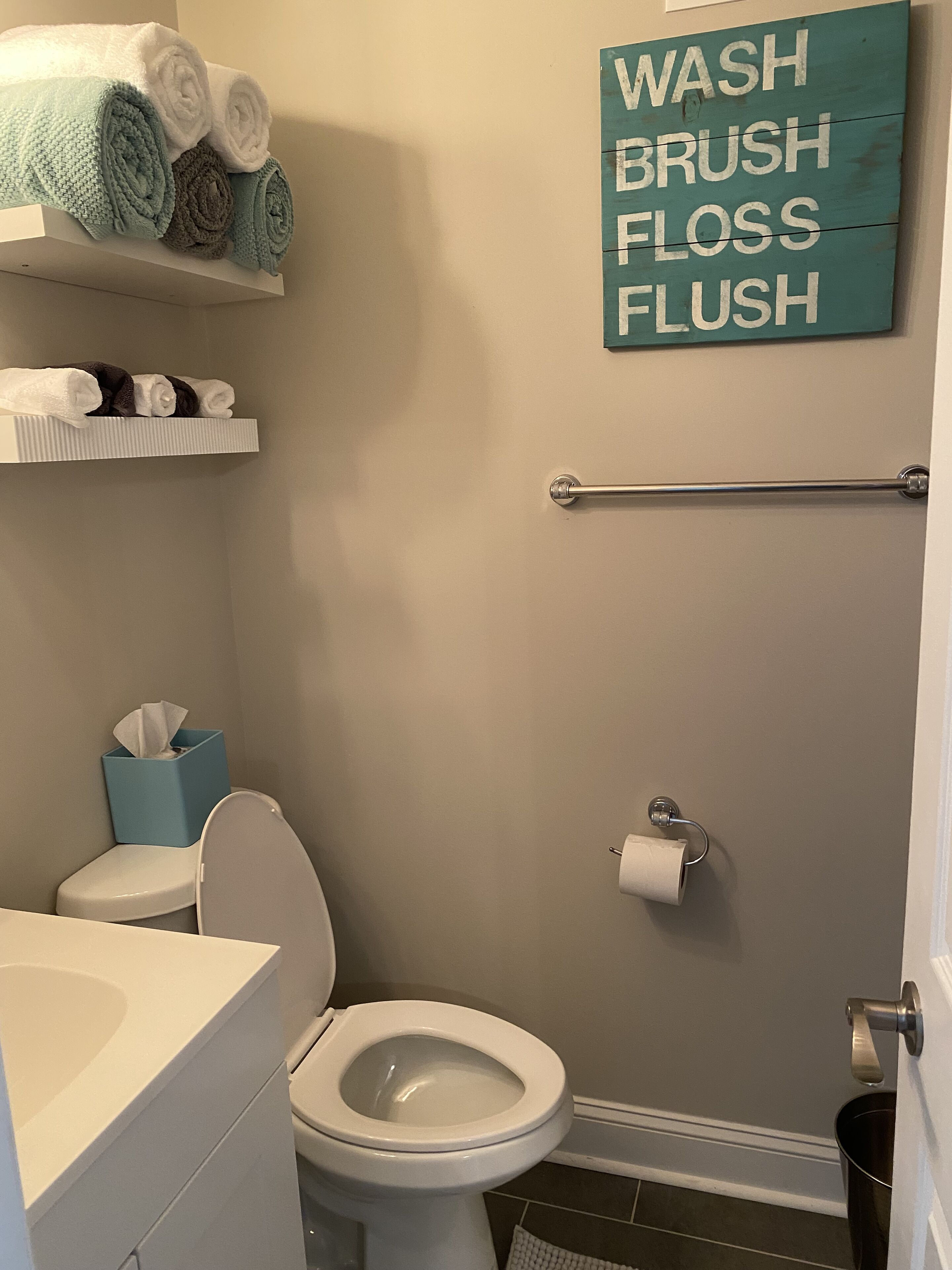 A bathroom with a toilet, a towel rack with towels, a toilet paper holder, and a sign that reads "WASH BRUSH FLOSS FLUSH".