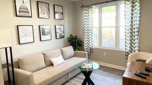 A living room with a beige sofa, a glass coffee table, and a wooden side table. There are framed pictures on the wall and a large window with curtains.