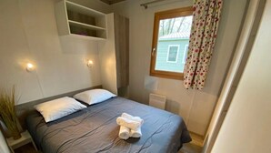 Un lit simple avec une couvre-lit grise et des oreillers blancs, une petite table de chevet, une étagère fixée au mur, une fenêtre avec des rideaux et un radiateur.