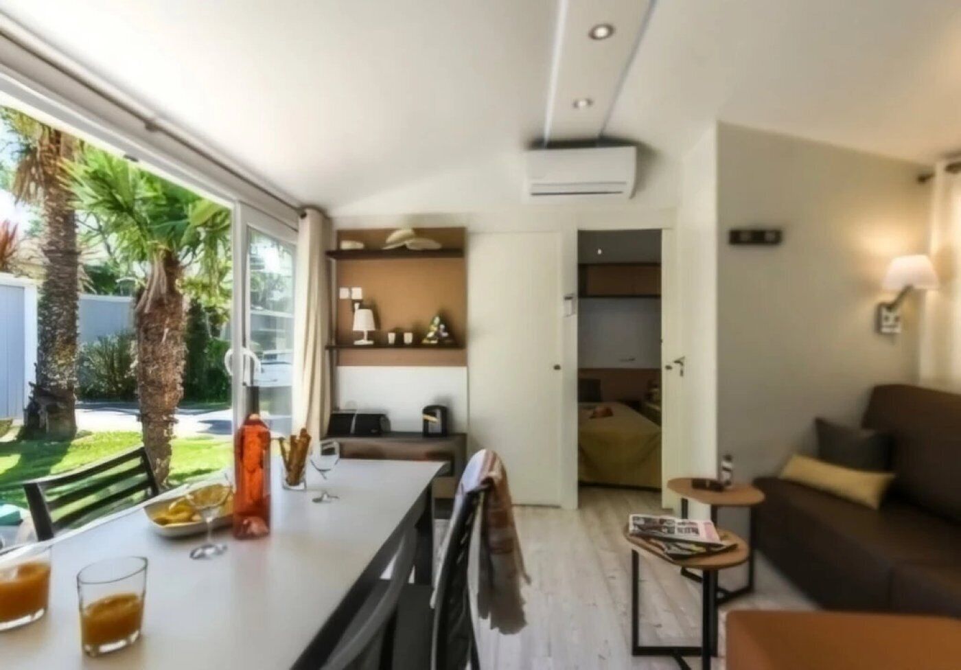 Ein modernes Wohnzimmer mit einer Couch, einem Couchtisch und einem Essbereich mit Tisch und Stühlen. Ein Fenster bietet Blick auf Palmen und es gibt einen Balkon.