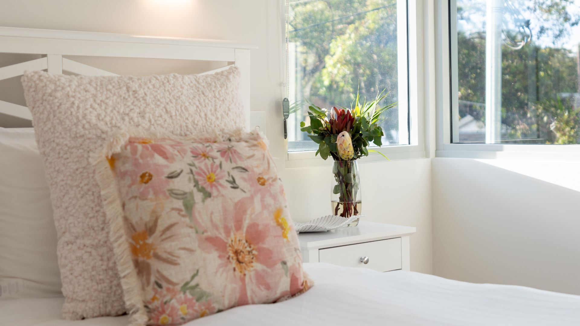A bedroom with a bed, a nightstand, a vase with flowers, and a window with a view of trees.