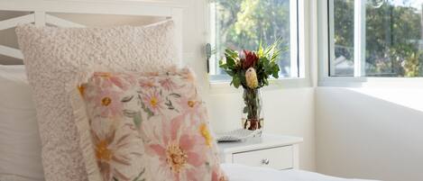 A bedroom with a bed, a nightstand, a vase with flowers, and a window with a view of trees.