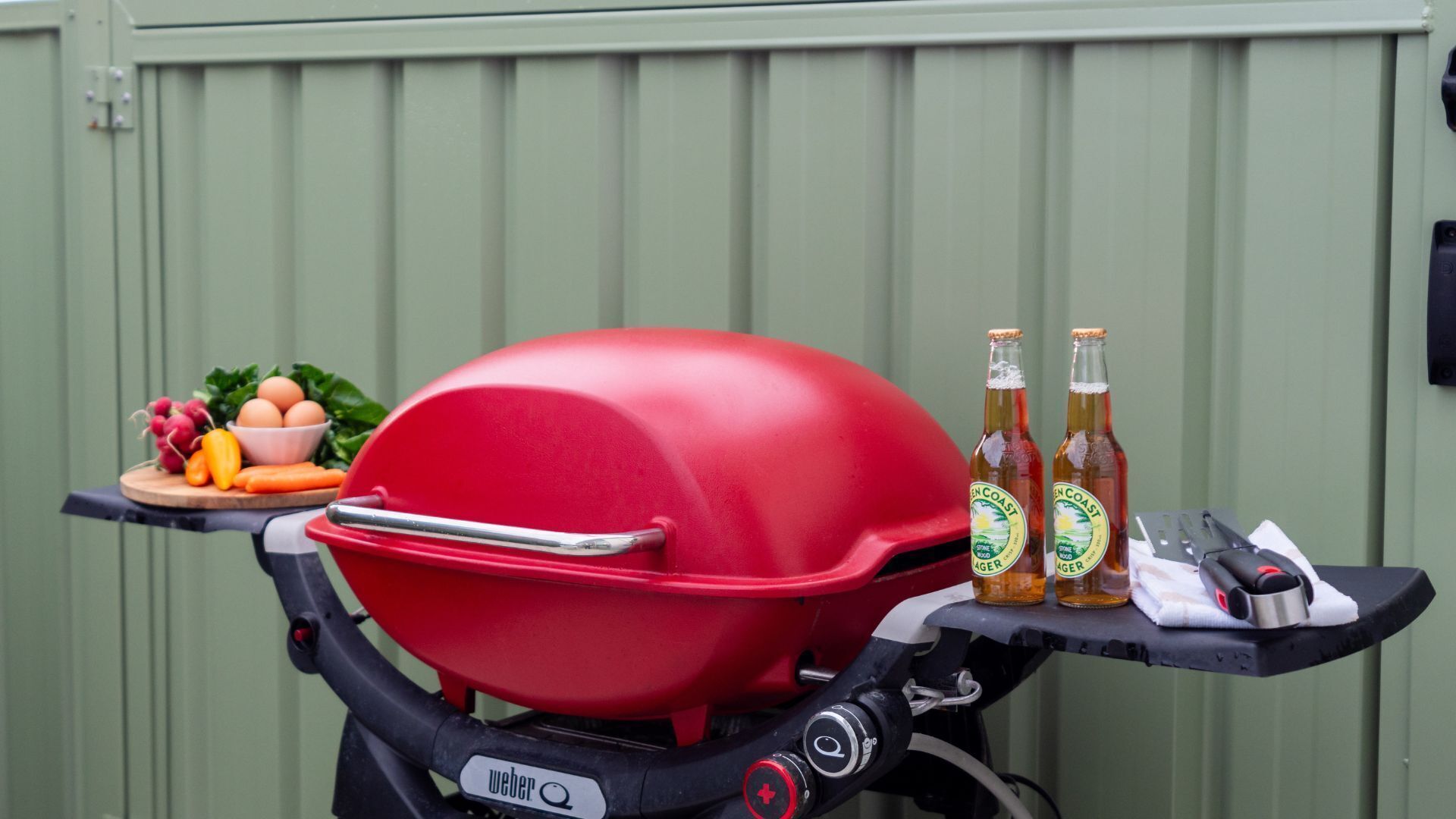 Una parrilla Weber roja con una bandeja de verduras y dos botellas de cerveza sobre una mesa plegable.