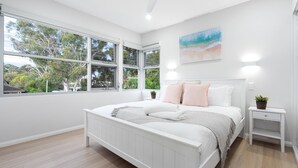 Une chambre avec un grand lit, une table de chevet, une lampe fixée au mur et une fenêtre donnant sur des arbres et une maison.