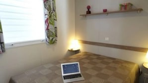 Ein Schlafzimmer mit einem Laptop auf dem Bett, einem Fenster mit Vorhängen, einer Nachttischlampe und einem Regal mit Büchern und einer Vase.