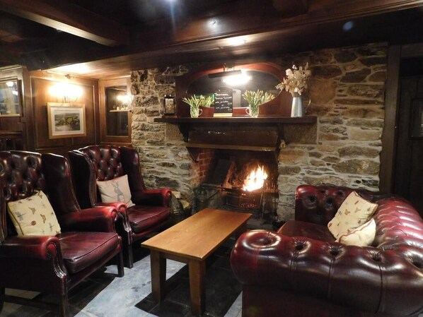 A cozy lounge area with leather sofas and armchairs, a wooden coffee table, and a stone fireplace with a lit fire.
