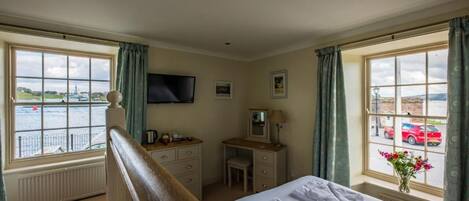 A hotel room with a bed, a desk, a television, and a view of the sea and a red car through the window.