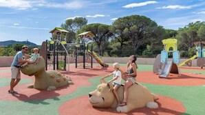 Un parque infantil con estructuras de juego, incluyendo un tobogán y zonas de escalada, rodeado de árboles y un cielo despejado.