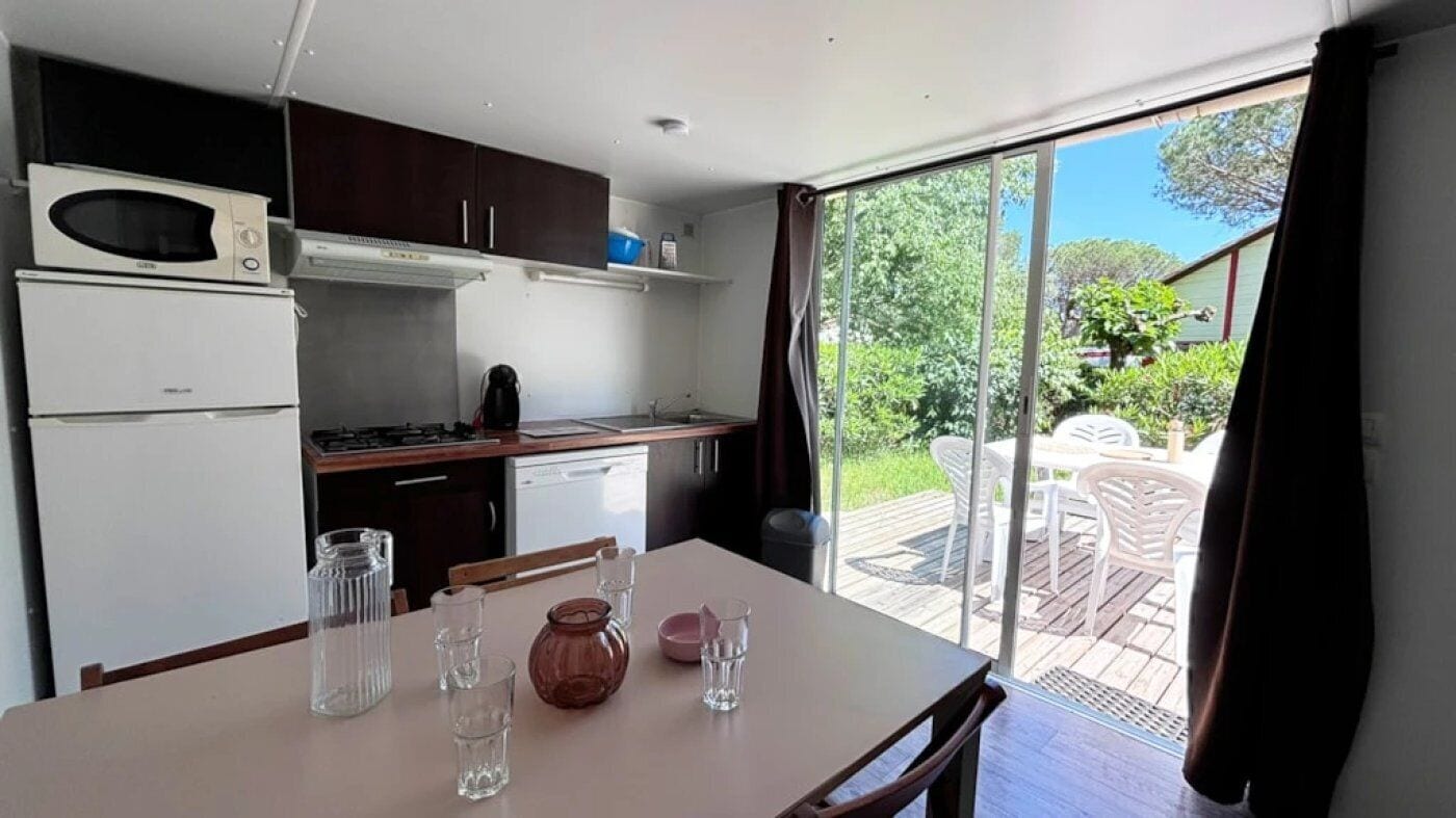 Une cuisine compacte équipée d’un four à micro-ondes, d’une cuisinière et d’un lave-vaisselle. On y trouve une table à manger avec des chaises, et une porte-fenêtre coulissante donnant sur un jardin.