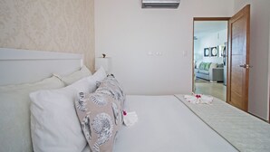 Una habitación de hotel con cama, mesita de noche, equipo de aire acondicionado y puerta que da a otra habitación.