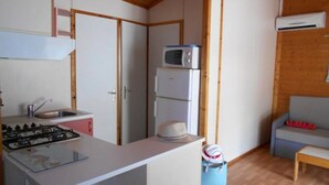 Pieni, käytännöllinen keittiö, jossa on kaasuliesi, pesuallas ja mikroaaltouuni jääkaapin päällä. Nurkassa on sininen roskakori ja pieni pöytä.