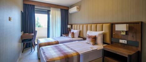 Habitación de hotel con dos camas, un escritorio y una silla. Cuenta con una ventana con cortinas, un equipo de aire acondicionado y una mesita de noche de madera.