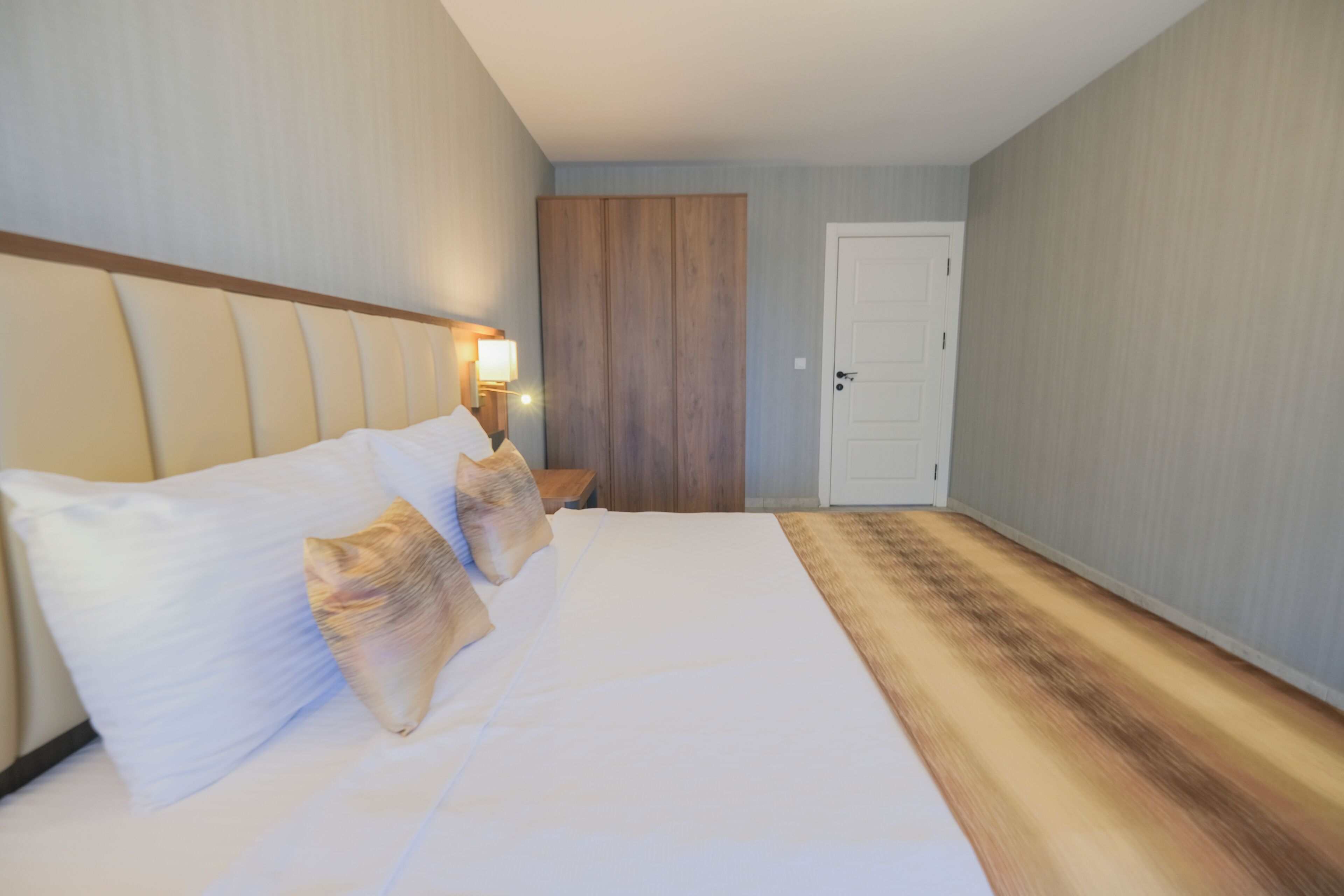 Una habitació d'hotel amb un llit gran, un armari de fusta, una porta blanca i una tauleta de nit amb una làmpada.