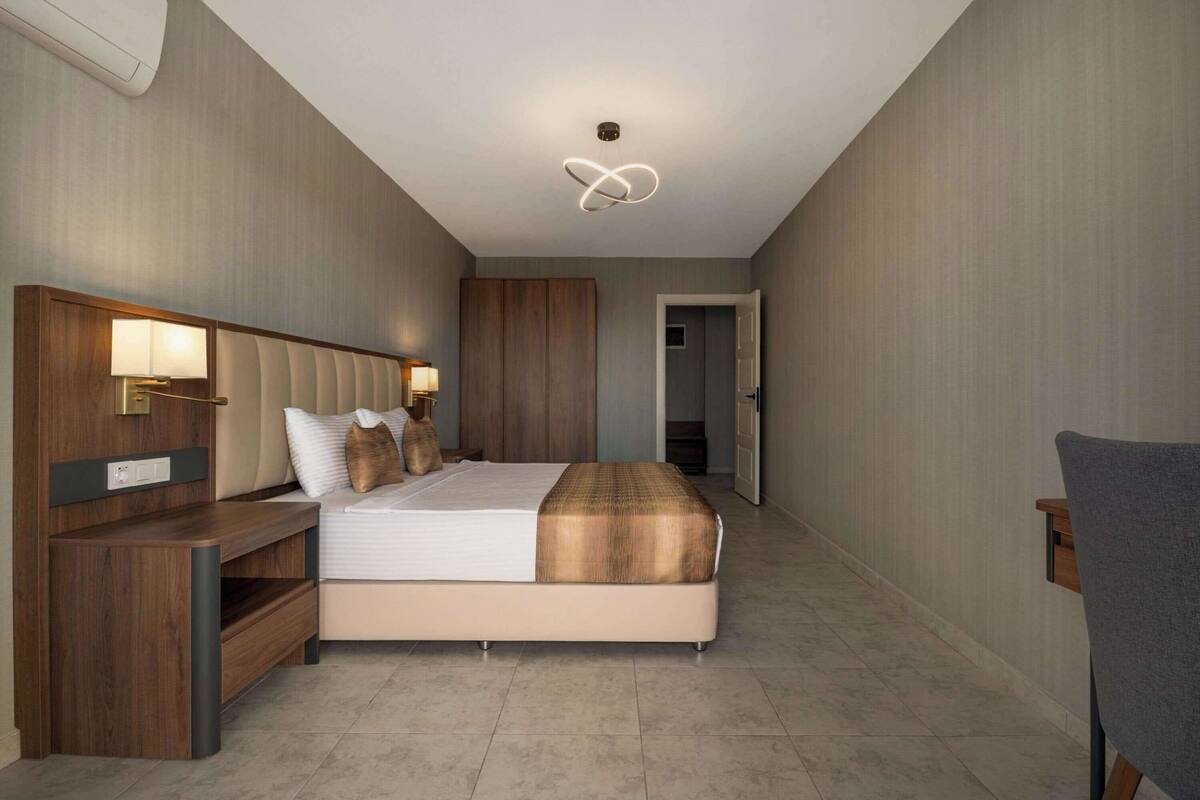 A modern hotel room with a large bed, bedside tables, a wardrobe, and a ceiling light fixture.
