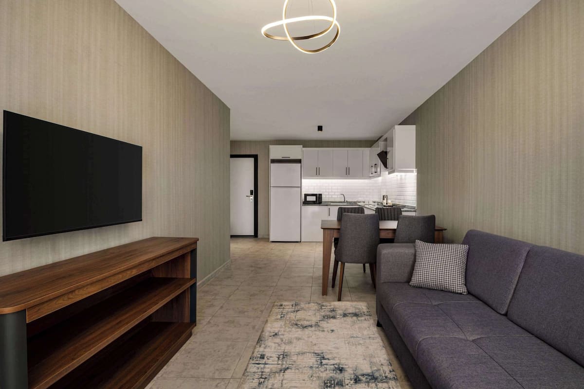 A modern living room with a flat-screen TV, a wooden entertainment unit, a grey sofa, and a kitchen in the background.