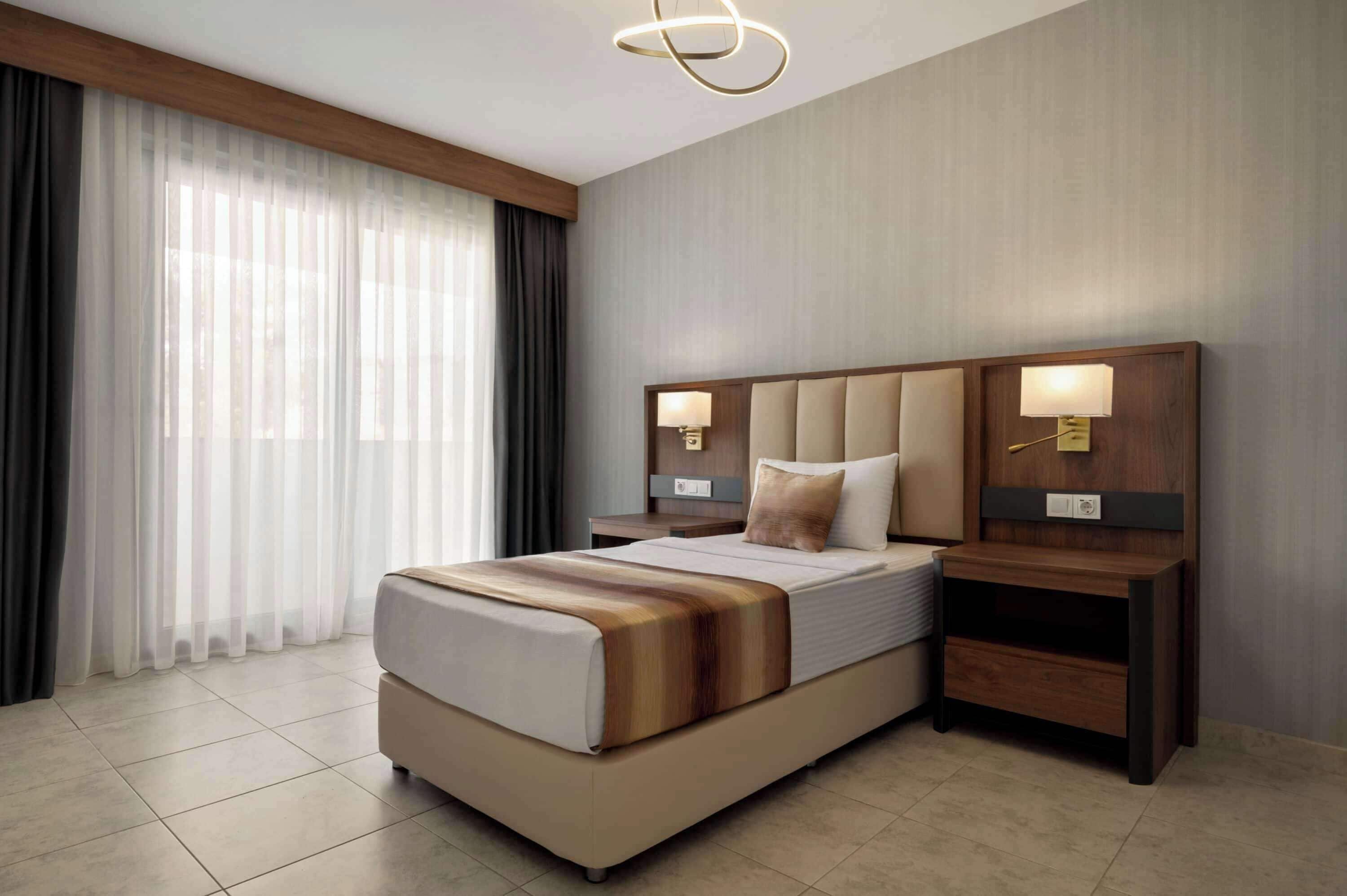 Una habitación de hotel moderna con una cama grande, cabecera de madera, dos mesitas de noche con lámparas y un ventanal con cortinas translúcidas.