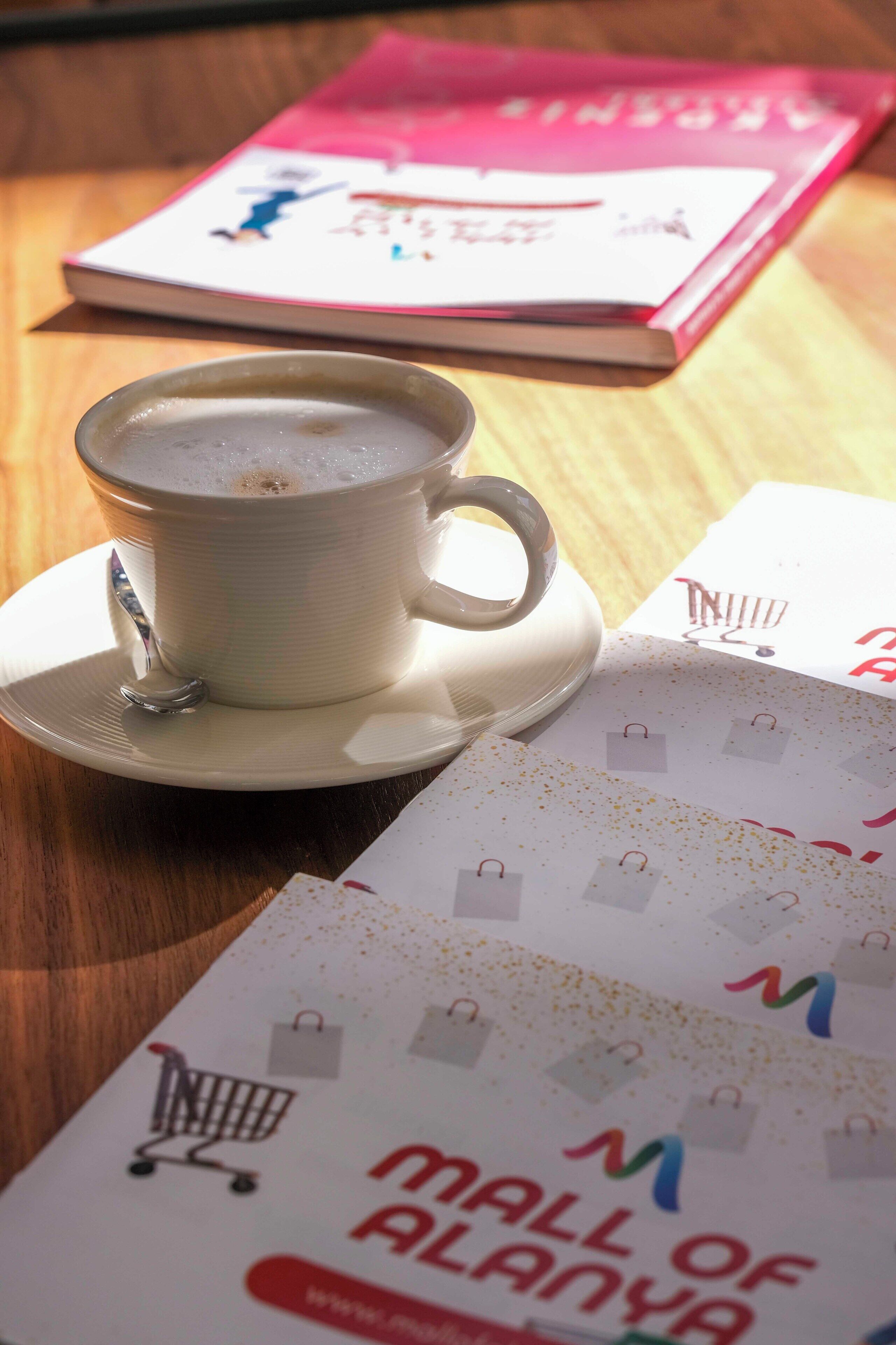 Uma chávena de café sobre um pires, uma mala de compras e um livro sobre uma mesa de madeira.