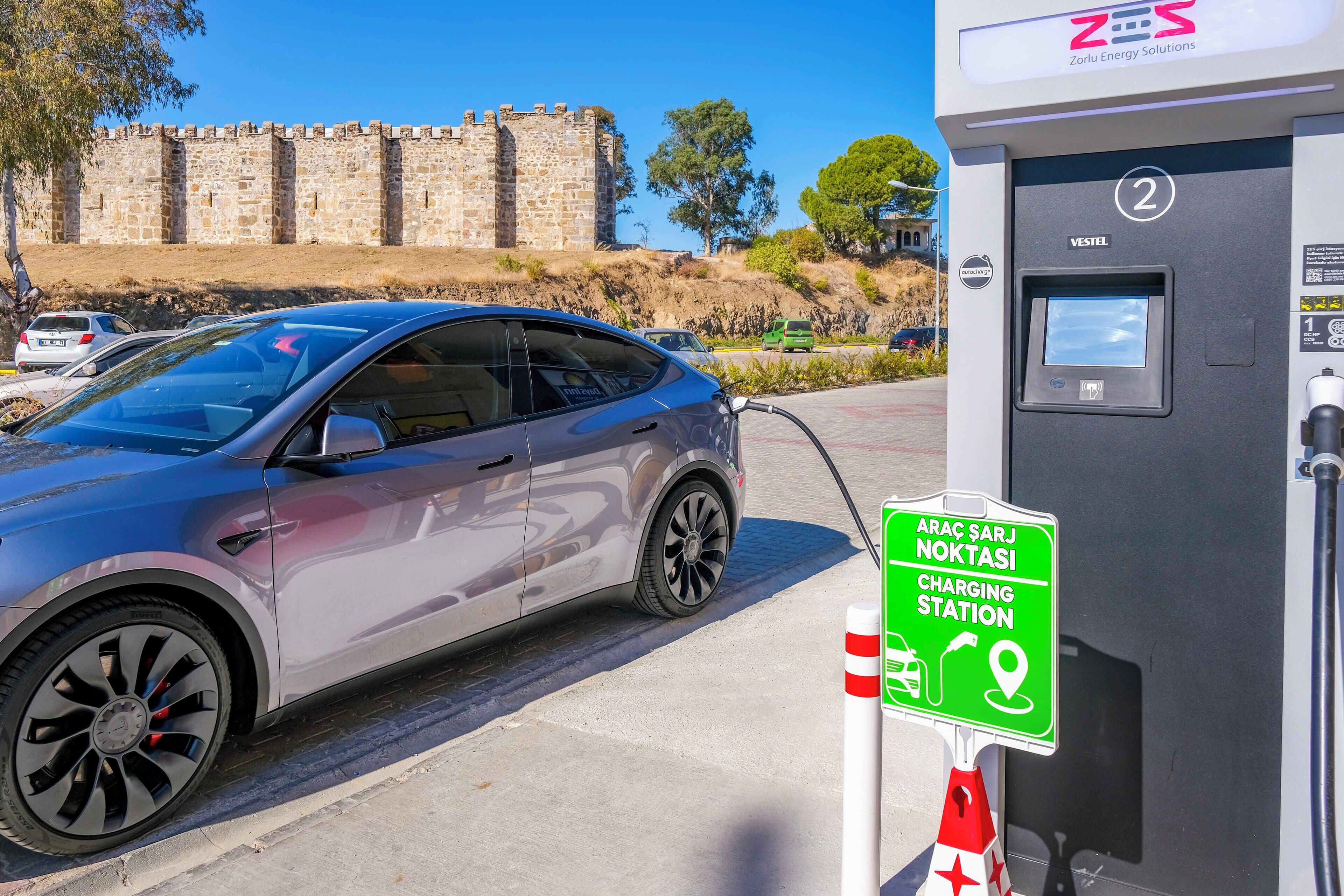 A charging station with a car connected to it, a sign indicating it's a charging station, and a stone wall in the background.