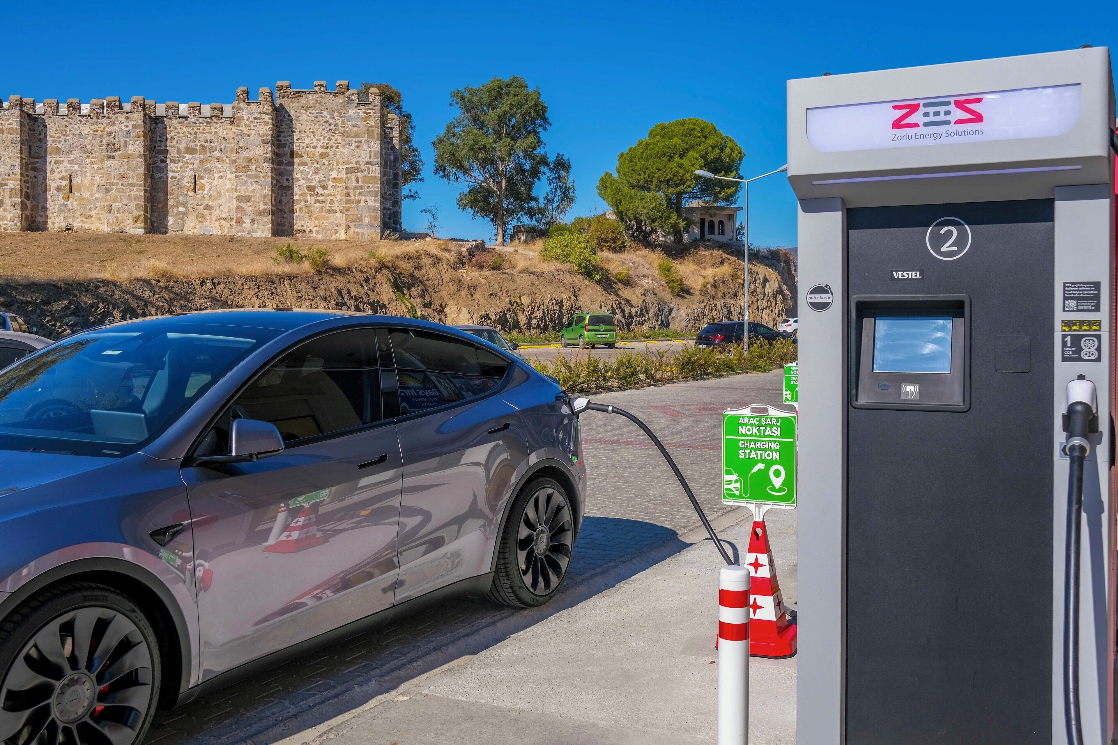 Una estación de carga con un automóvil estacionado cerca, un muro de piedra y árboles al fondo.
