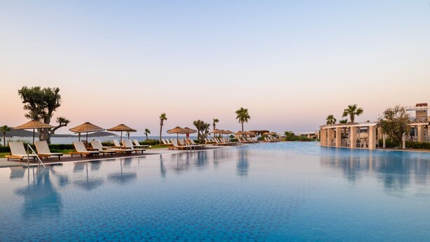 A pool with lounge chairs and umbrellas, palm trees, and a building in the background.
