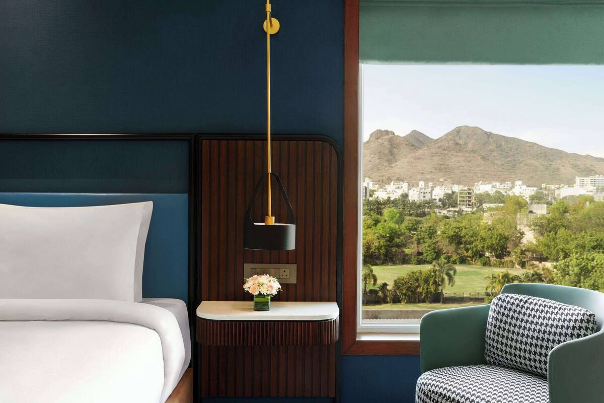 Hotelkamer met een bed, een tafeltje met een bloemenvaas, een hanglamp, een stoel en een raam met uitzicht op bergen en een stad.