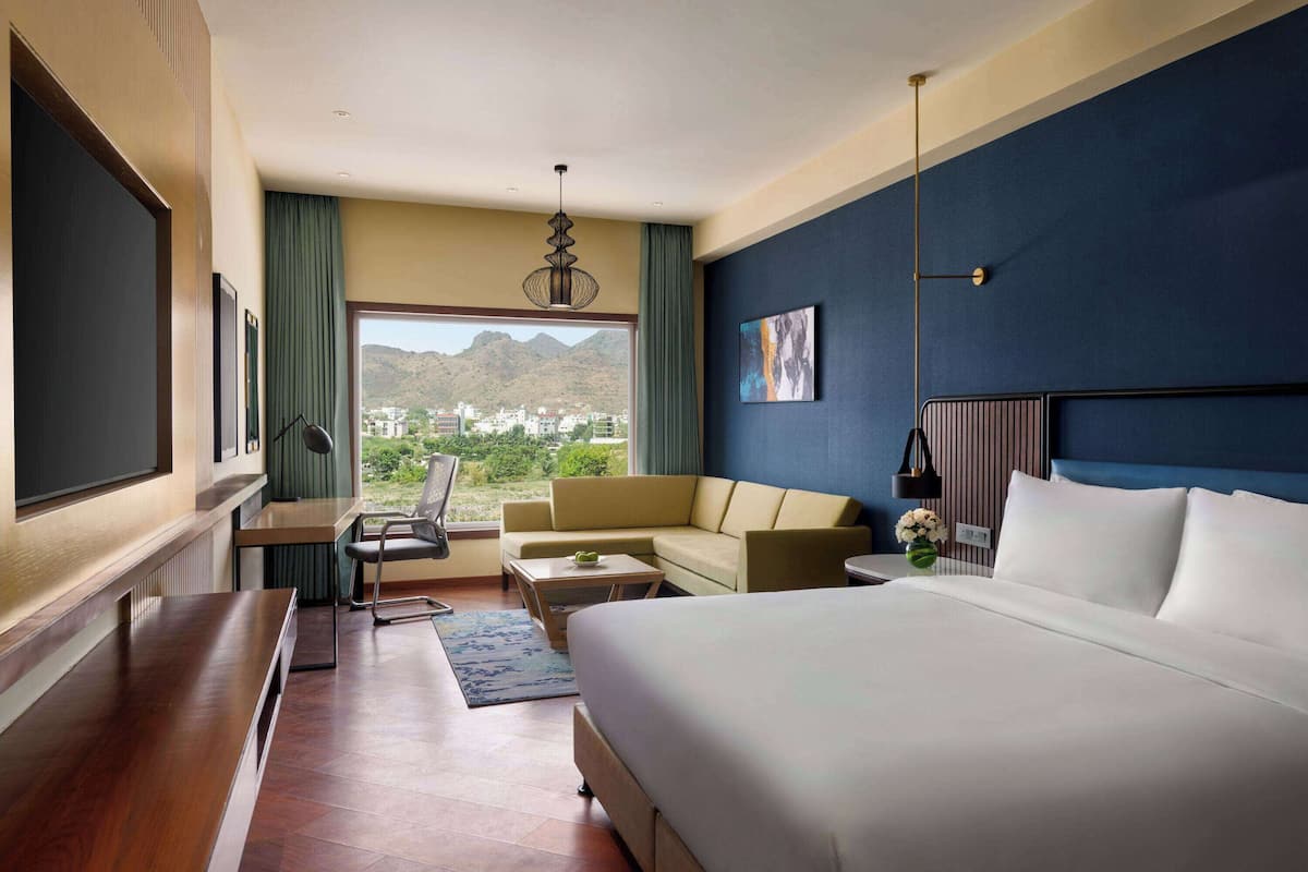 Een hotelkamer met een groot bed, een bank, een bureau en een stoel. Door het raam is er uitzicht op bergen en gebouwen.