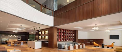 A spacious lobby with a high ceiling, wooden paneling, a reception desk, seating areas, and a large bookshelf.