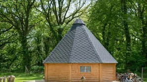 A wooden log cabin with a conical roof, surrounded by lush greenery and a gravel path leading to it.