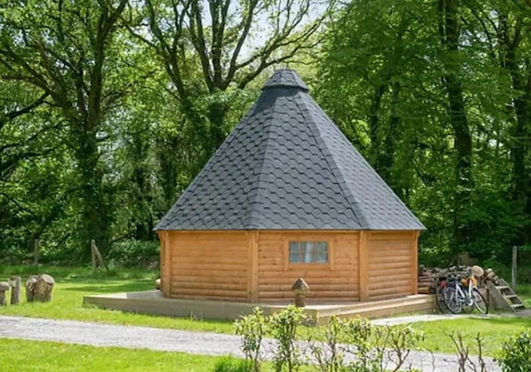 A wooden log cabin with a conical roof, surrounded by lush greenery and a gravel path leading to it.