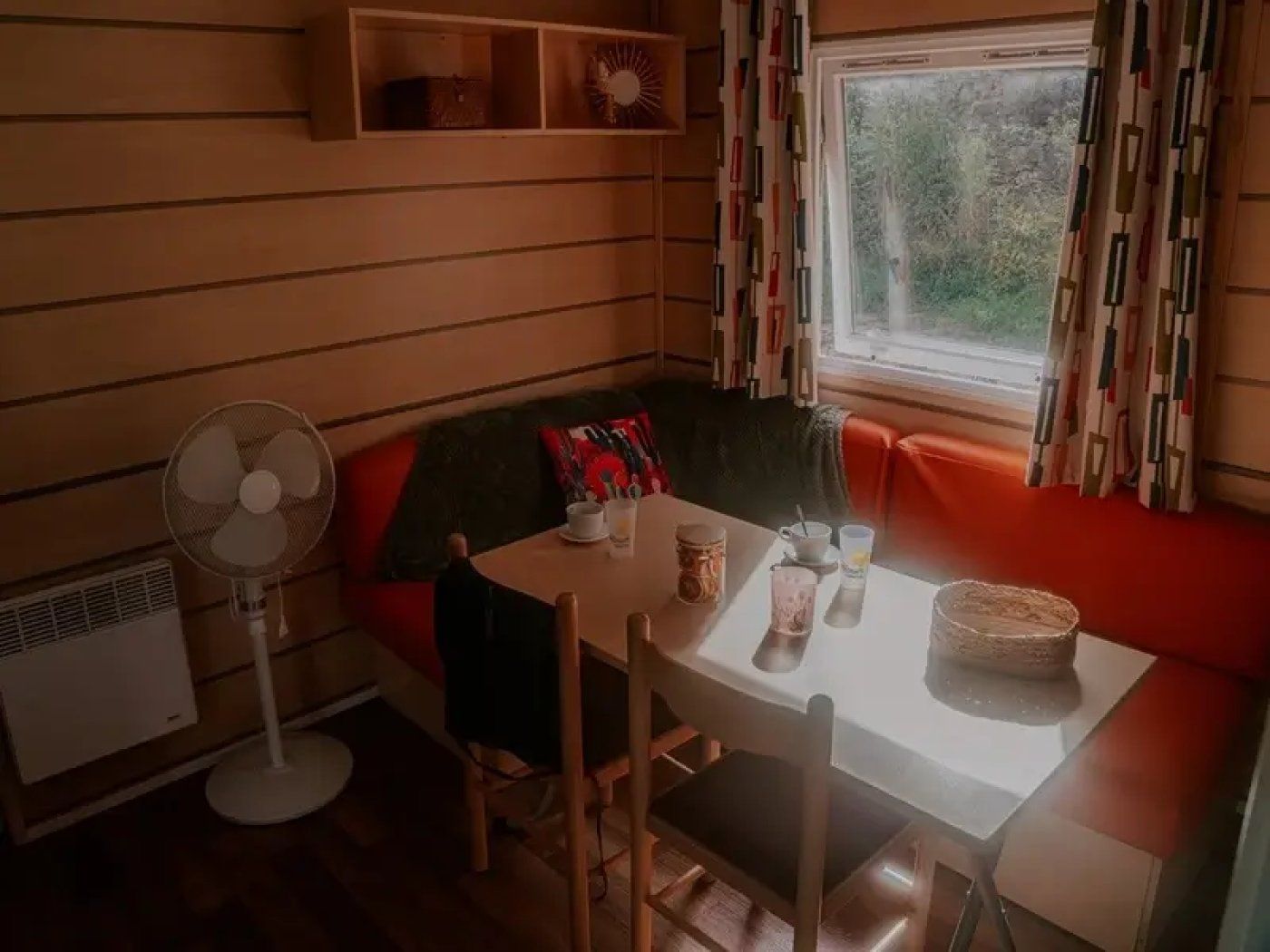 A cozy room with a wooden interior, a table with cups and a fan, and a window with curtains.
