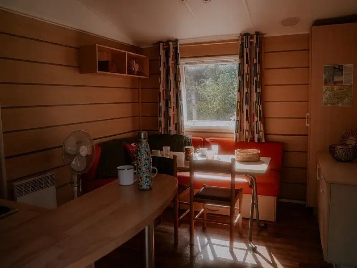 A cozy room with a wooden interior, a window with curtains, a table with chairs, and a small shelf with items.