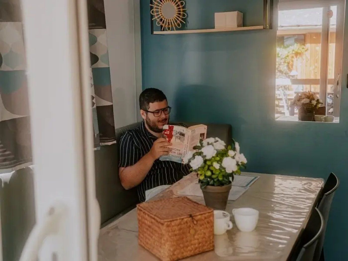 A person reading a book at a table with a potted plant and a basket.