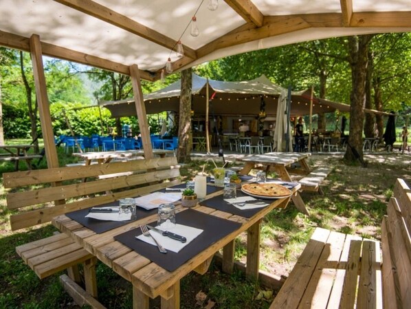 Un área de comedor al aire libre con mesas y bancos de madera, rodeada de árboles y cubierta por una pérgola.