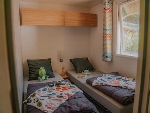 A compact room with two beds, each with a pillow and a blanket. There is a window with a curtain, a wall-mounted light, and a small table with a vase.
