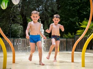 Dos niños jugando en una fuente de agua, con una cerca de madera y follaje al fondo.