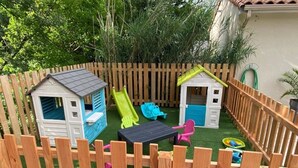 Een omheinde achtertuin met twee speeltuintjes, een glijbaan en een kleine tafel met stoelen.