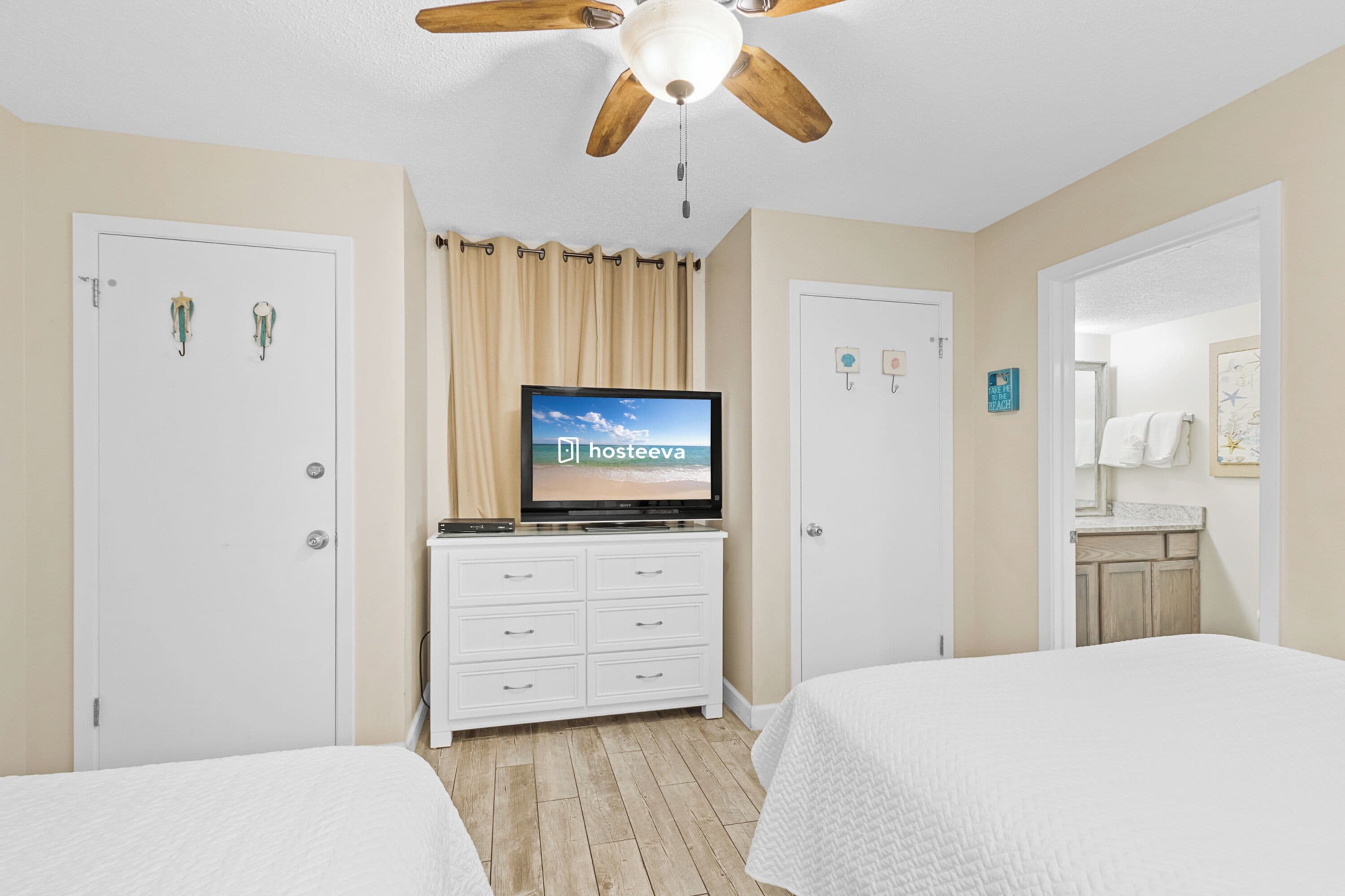A hotel room with two beds, a ceiling fan, a TV on a dresser, and a bathroom visible through an open door.