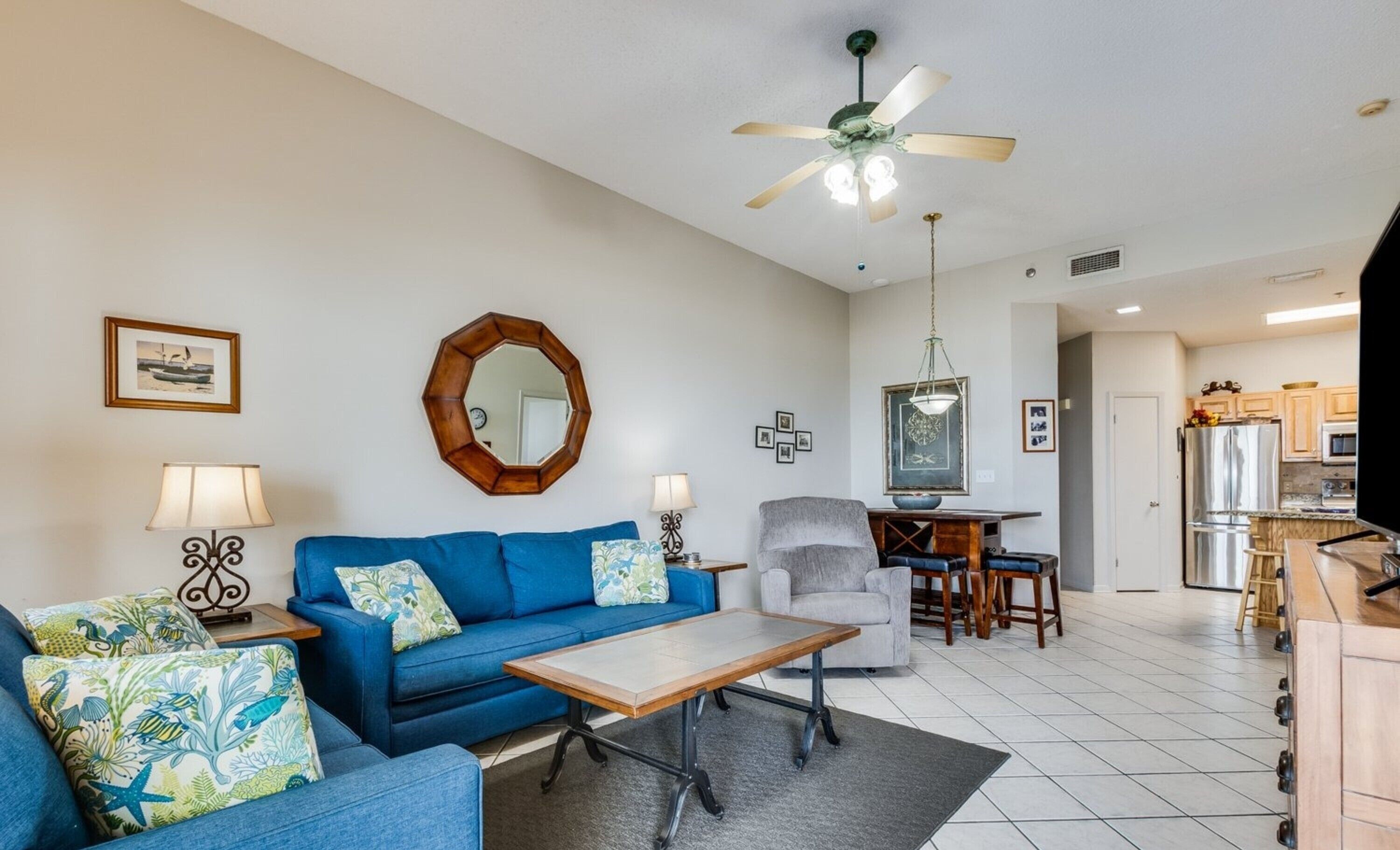 A living room with a blue sofa, a wooden coffee table, a grey armchair, and a dining area with a wooden table and chairs.
