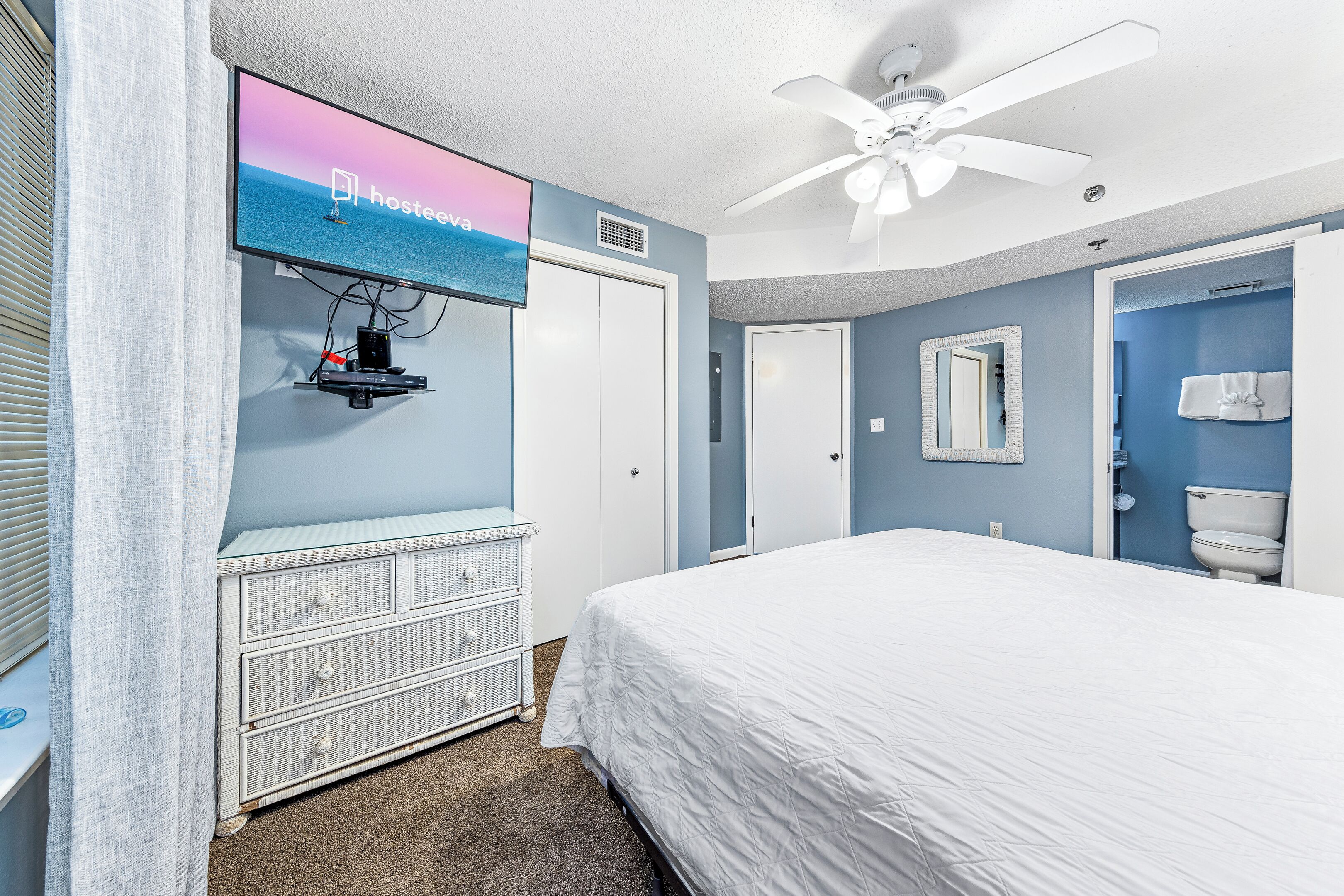 A bedroom with a bed, a television mounted on the wall, a ceiling fan, and a bathroom with a toilet and towel hanging on the wall.