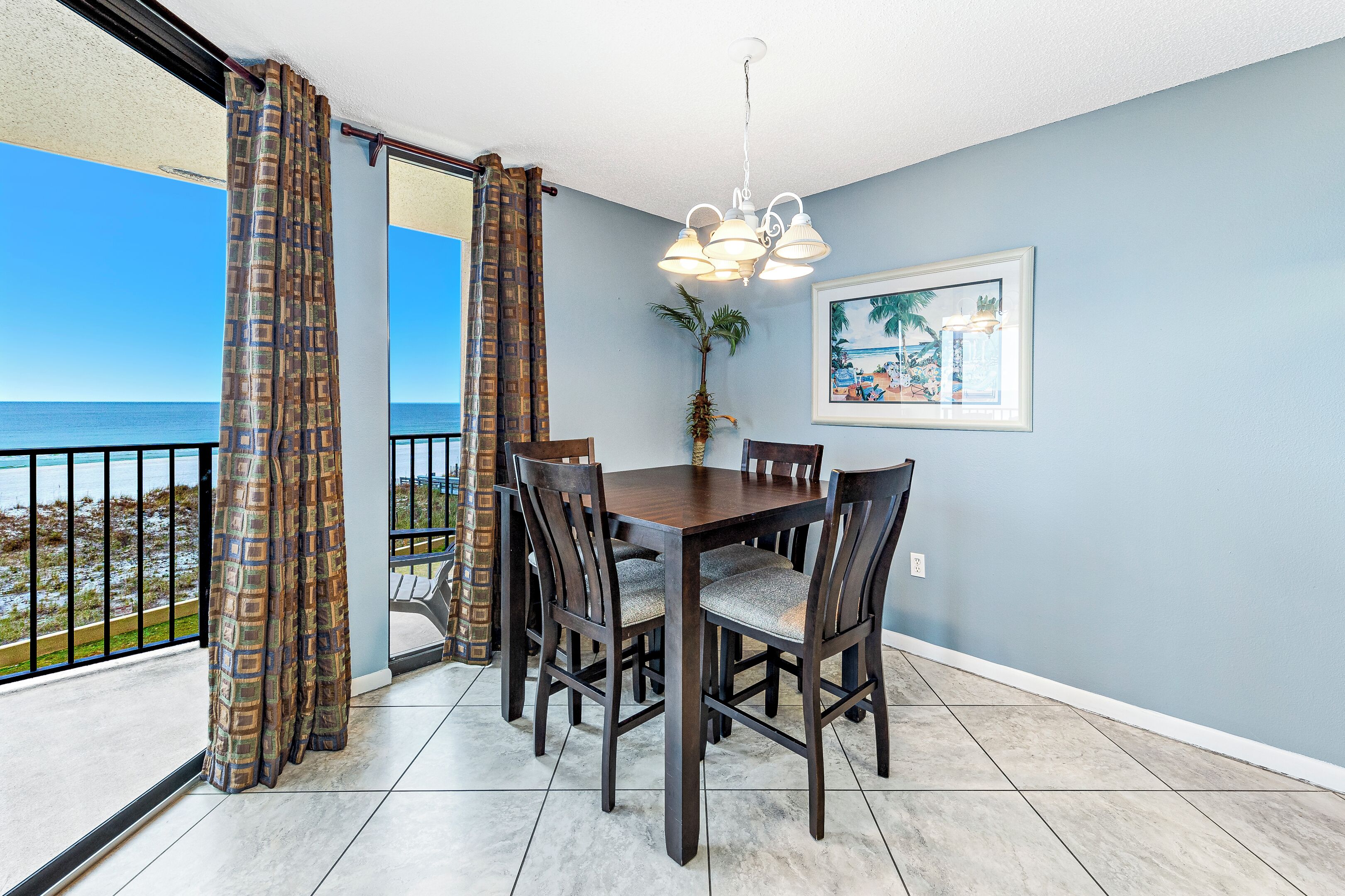 A dining area with a table and chairs, a potted plant, and a view of the ocean through a balcony with patterned curtains.