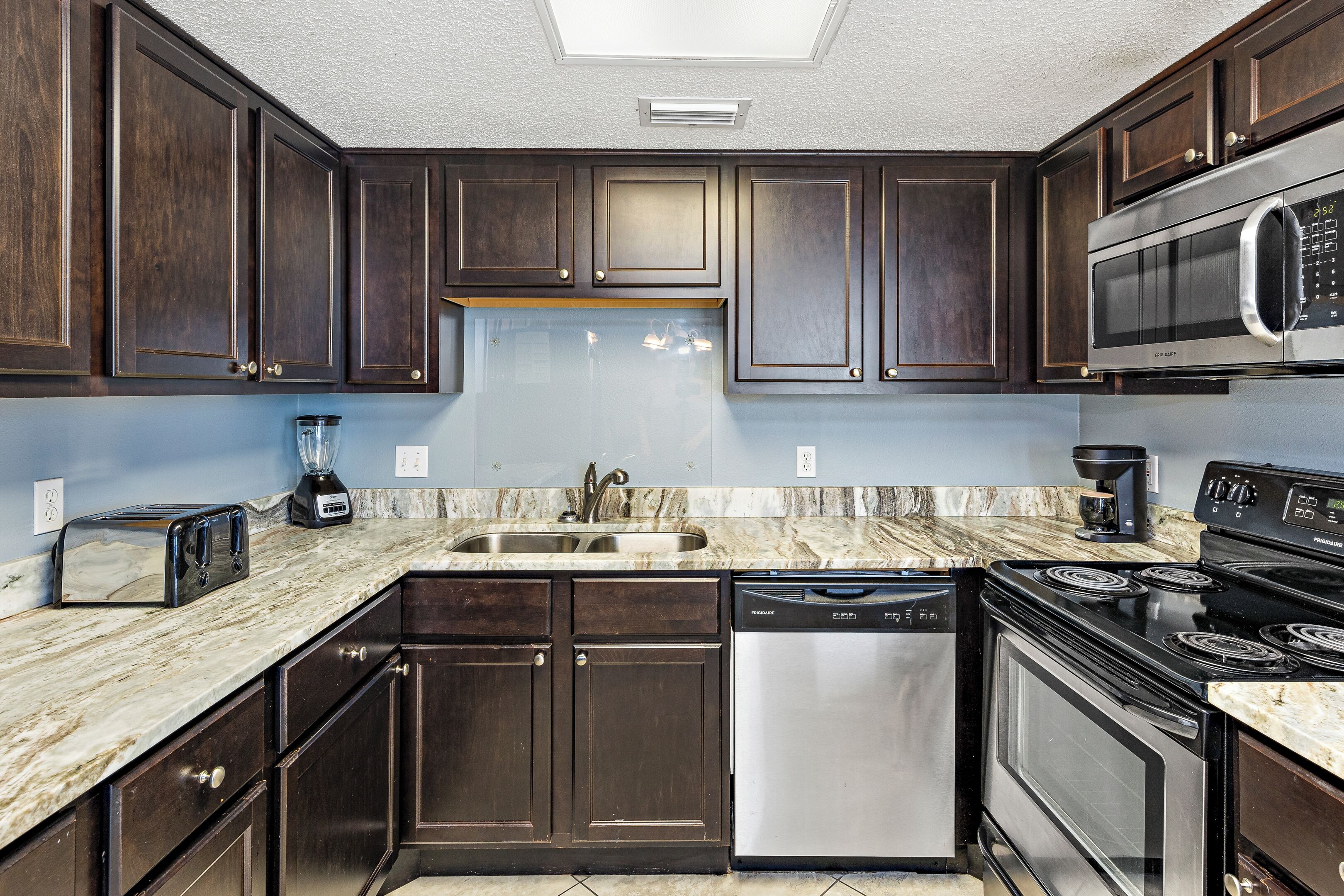 A modern kitchen with dark wooden cabinets, a stainless steel dishwasher, a built-in microwave, a black stove, and a marble countertop.