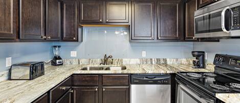 A modern kitchen with dark wooden cabinets, a stainless steel dishwasher, a built-in microwave, a black stove, and a marble countertop.