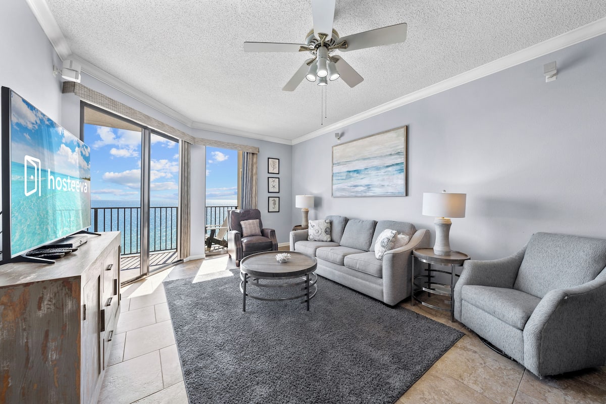 Un salón amplio con un sofá gris, una mesa de centro y un televisor. Hay un balcón con vistas al mar.